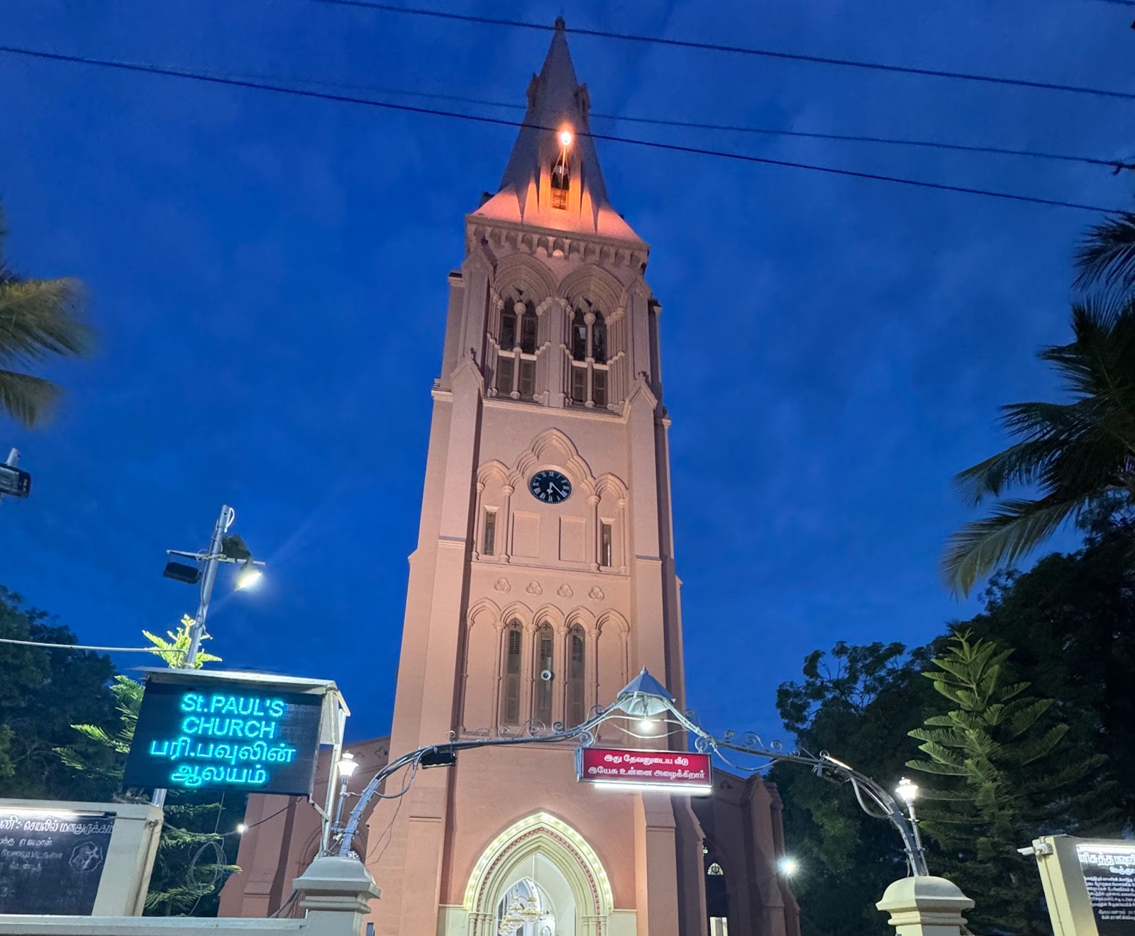 The tower and spire of St. Paul&rsquo;s Church, Meignanapuram, lit at dusk against a deep blue sky, with palms on either side and an illuminated sign reading St. Paul&rsquo;s Church in English and Tamil.